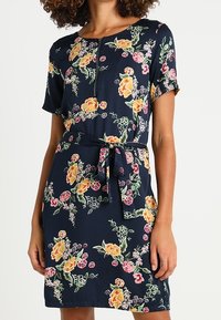 Robe florale marine à manches courtes, avec un détail de bouton et une taille nouée. Motifs floraux jaunes, roses et verts sur un fond sombre.
