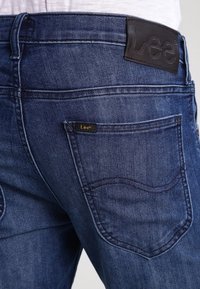 Mörkblå denimjeans med en läderetikett i midjan som har "Lee". Blekad textur och kurviga sömdetaljer på bakfickorna.