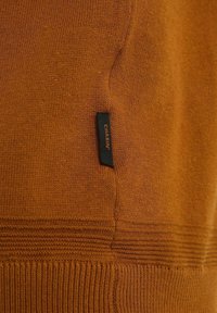 Gros plan sur un tissu marron en tricot avec une étiquette rectangulaire noire portant l'inscription « CHASIN' » cousue près de l'ourlet inférieur.