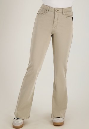 Beige hoge taille rechte broek gedragen met witte sneakers door een staande persoon tegen een effen achtergrond.