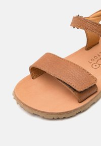 Bruna lädersandaler med två breda justerbara remmar, en beige fotbädd och en texturerad gummisula. Sömnadsdetaljer synliga på remmarna.