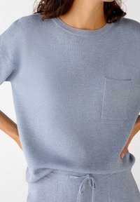 Nainen, joka on pukeutunut vaaleansinisestä neulotusta lyhythihaisesta paidasta, jossa on etutasku, ja samansävyisistä nyöritettävistä housuista, kädet lantiolla.