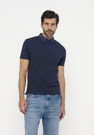 Uomo che indossa una polo blu navy a maniche corte con colletto decorato e jeans blu su sfondo bianco.