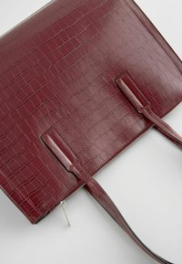 Sac cabas en cuir embossé crocodile rouge foncé avec double poignée et une fermeture éclair argentée le long du bord supérieur.