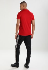 Rotes Polohemd mit Kragen, kombiniert mit schwarzen Sporthosen mit weißen Seitenstreifen und schwarzen Sportschuhen.