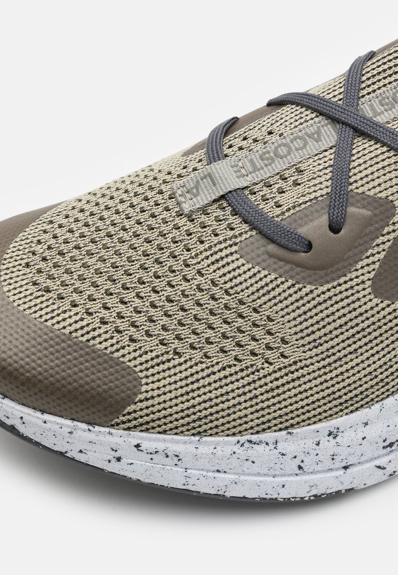 Zapatilla deportiva ligera con una parte superior de malla transpirable en tonos beige y acentos en gris oscuro, cordones redondos y una suela blanca con manchas.