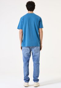 Türkises Baumwoll-T-Shirt mit Rundhalsausschnitt, kombiniert mit hellblauen geraden Jeans und beigen Sneakers, von hinten betrachtet.