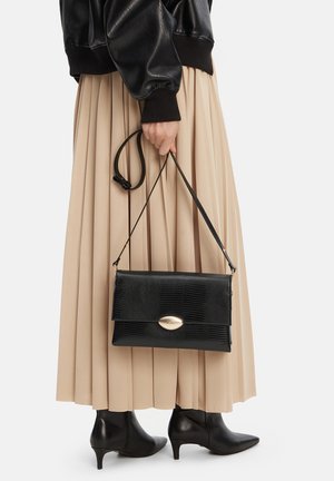 Frau in beige plissiertem Rock und schwarzer Lederjacke, die eine schwarze, strukturierte Handtasche mit goldenem Verschluss hält, dazu schwarze Stiefeletten mit Absatz trägt.