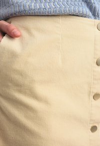 Jupe en velours côtelé beige avec des côtes verticales, présentant une texture lisse et des boutons dorés sur le côté. Main tenant le tissu.