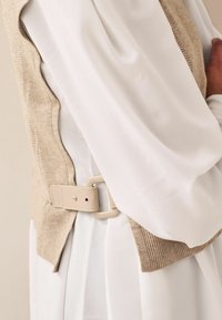Béžová pletená vesta s ribbed texturou, vrstvená přes bílou blůzu. Obsahuje světle béžový pás s obdélníkovou sponou. Elegantní a moderní design.