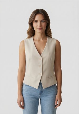 Donna con capelli castani mossi, indossa un gilet beige senza maniche con bottoni e jeans azzurri, in piedi su uno sfondo grigio chiaro uniforme.