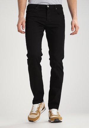 Homme portant un jean noir slim et des baskets blanches, marron et beige, debout devant un fond clair uni.