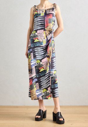 Woman wearing a colorful, graphic print midi dress with spaghetti straps and black platform heeled sandals, standing on wooden floor.
