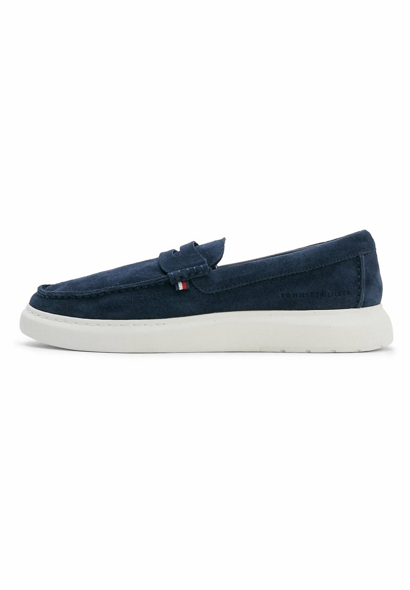 Mocassin en daim bleu marine avec semelle blanche en caoutchouc, couture style mocassin, et petit détail en tissu rouge, blanc et bleu sur le côté.