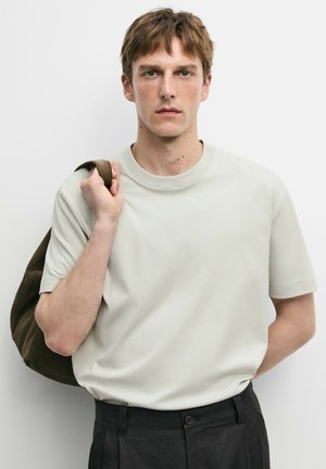 Junger Mann mit kurzen braunen Haaren, der ein hellbeiges T-Shirt, schwarze Hose trägt und eine braune Tasche über der Schulter trägt.