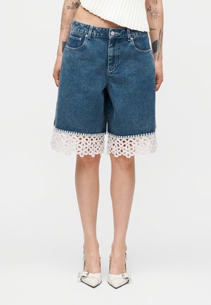 Jeansshorts mit weißem floralen Spitzenbesatz am Saum, getragen von einer tätowierten Person mit weißen spitzen Absätzen und einem kurzen weißen Oberteil.