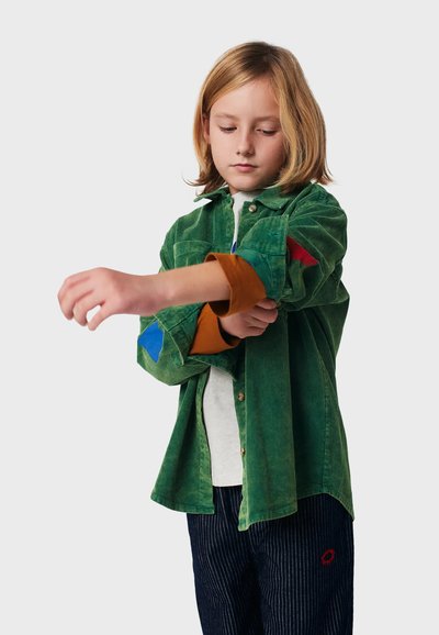Kind mit schulterlangem blonden Haar, der den Ärmel eines grünen Cordhemds mit bunten Patches hochrollt, trägt gestreifte Hosen.