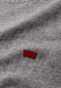 Tissu tricoté gris avec un petit patch de logo rouge cousu près du centre, de forme rectangulaire avec un bord inférieur courbé.