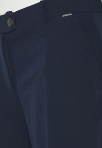 Granatowe spodnie wykonane z gładkiego materiału, z zapięciem na guzik, kieszenią boczną oraz subtelnym akcentem z logo w pobliżu pasa.