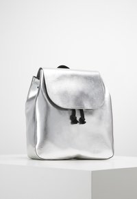 Sac à dos métallique argenté de forme arrondie, rabat à rabat et fermeture par cordon noir. Texture lisse avec un éclat subtil.