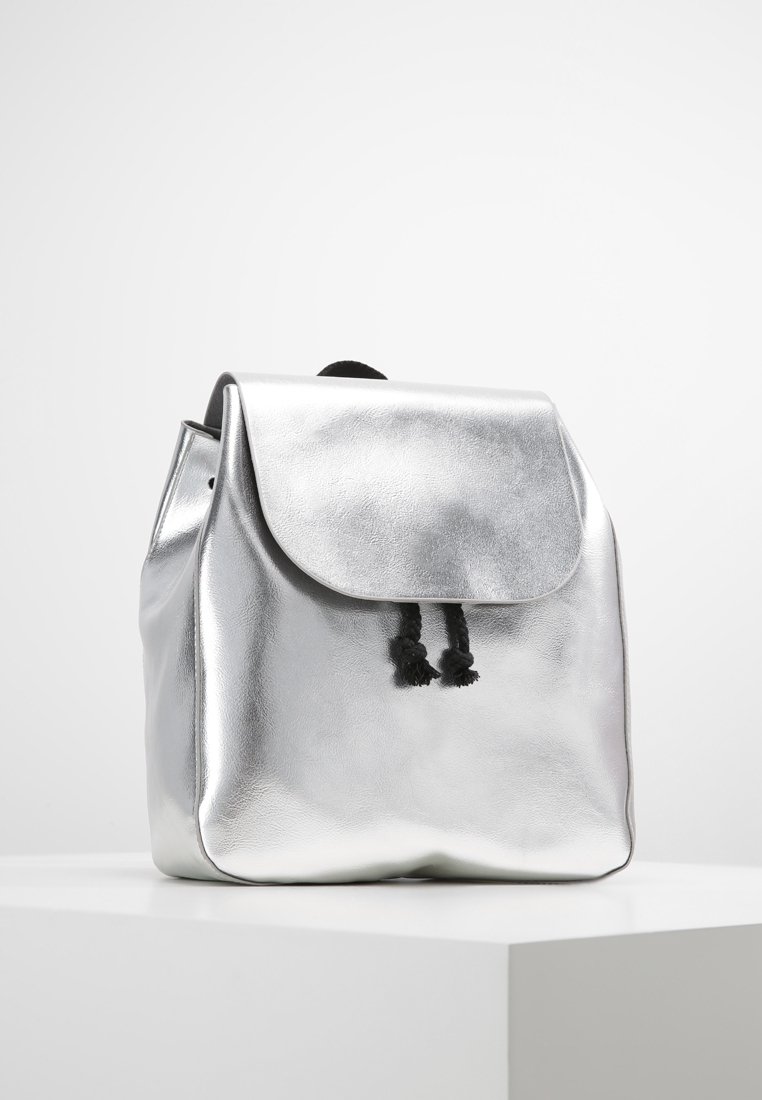 Sac à dos métallique argenté de forme arrondie, rabat à rabat et fermeture par cordon noir. Texture lisse avec un éclat subtil.