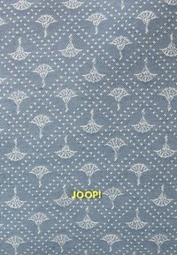 Denimkangas vaaleansinisen taustan kanssa, jossa on valkoisia tuulettimen kuvastoja ja koristeellisia pisteitä; "JOOP!" logo kirkkaan keltaisena.