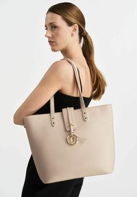 Beżowa skórzana torba typu tote o zdefiniowanym kształcie, z dwoma d�ługimi uchwytami, złotym akcentem w formie pierścienia oraz górnym zapięciem z detalami w postaci paska.