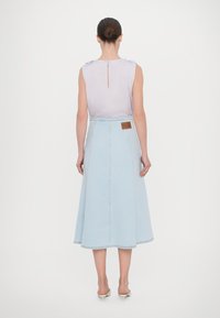 Kvinna med håret i en knut, klädd i en ärmlös lavendelfärgad topp och en ljusblå denim midi-kjol med beige kitten heels, stående vänd bort.