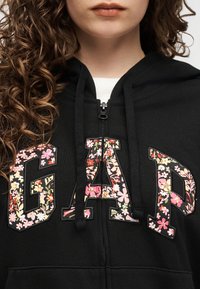 Γυναίκα με σγουρά μαλλιά που φοράει μαύρο φούτερ με φερμουάρ και μεγάλα γράμματα με λουλουδάτο σχέδιο που σχηματίζουν τη λέξη "GAP" στο στήθος.