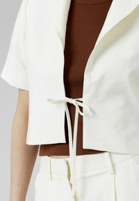 Veste blanche cintrée à manches courtes, avec une fermeture à lacer à l'avant, superposée à un débardeur marron à côtes.