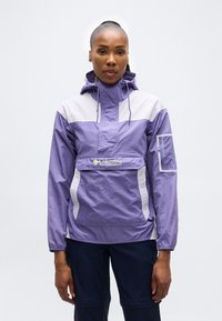 Γυναίκα με μπουφάν αδιάβροχο Columbia Sportswear σε μωβ και λευκό χρώμα με κουκούλα και σκούρο μπλε παντελόνι, στέκεται μπροστά σε απλό λευκό φόντο.