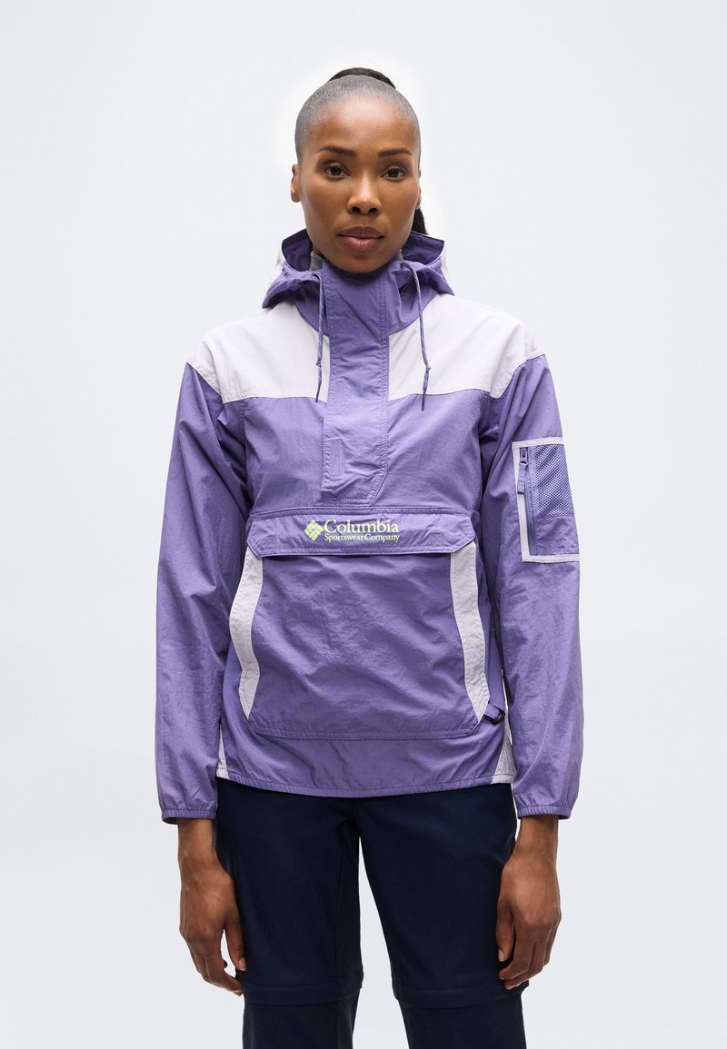 Γυναίκα με μπουφάν αδιάβροχο Columbia Sportswear σε μωβ και λευκό χρώμα με κουκούλα και σκούρο μπλε παντελόνι, στέκεται μπροστά σε απλό λευκό φόντο.