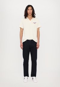 Kortegalm, off-white polo met een kraag en een rood logo, gecombineerd met donkerblauwe spijkerbroeken en witte sneakers.
