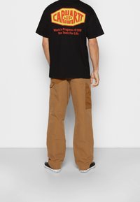 Muž oblečený v černém tričku Carhartt se žlutým a červeným logem a nápisem, hnědých cargo kalhotách a černých teniskách Converse, stojí proti bílé zdi.