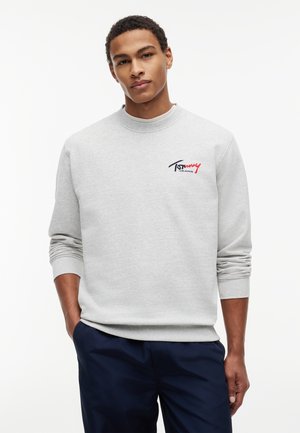 Młody mężczyzna w jasnoszarej bluzie Tommy Jeans z logo na piersi i ciemnoniebieskich spodniach, stojący z rękami w kieszeniach.