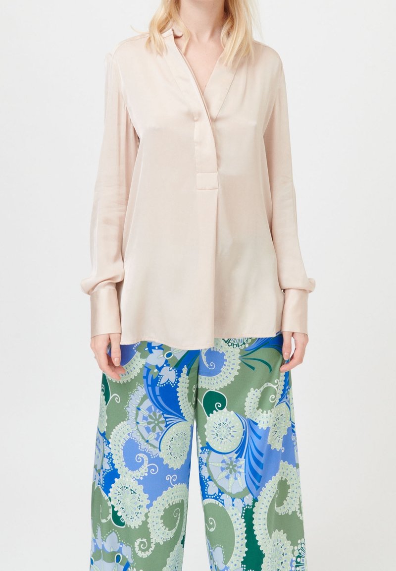 Vrouw in een losse lichtroze satijnen blouse en brede broek met een blauw, groen en wit paisley bloemenpatroon.