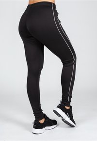 Zwarte leggings van rekbaar materiaal met een gladde textuur, voorzien van witte zijkanten en een verstelbare tailleband. Gecombineerd met zwarte sneakers.