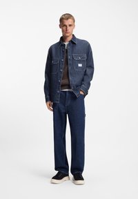 Chaqueta de mezclilla azul oscuro con botones al frente y dos bolsillos en el pecho, combinada con un suéter marrón de punto y pantalones vaqueros de corte holgado, acompañados de zapatillas negras.