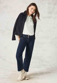 Veste noire texturée portée sur une chemise blanche, associée à un jean bleu foncé et des baskets beiges. Comprend un col et des boutons sur le jean.