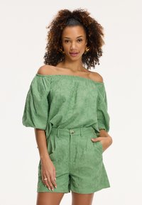 Blusa verde a spalle scoperte con motivo floreale testurizzato, abbinata a pantaloni corti a vita alta con chiusura a bottone e tasche laterali.