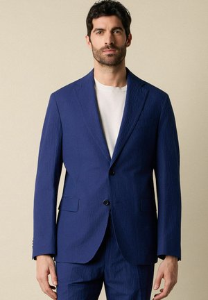 Man met donker haar en baard, gekleed in een gestructureerd blauw colbert en bijpassende broek over een wit shirt met ronde hals, staand tegen een beige achtergrond.