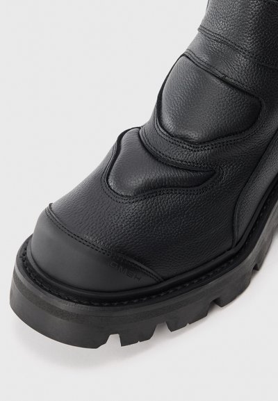 Botas de cuero negro con acabado texturizado, punta redonda, costuras distintivas y suela robusta con relieve. Logo de la marca visible en el talón.