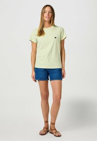 T-shirt di cotone verde chiaro con un piccolo logo blu navy, abbinato a pantaloni corti in denim blu scuro. Il modello indossa sandali marroni con cinturini.