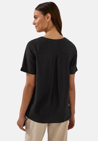 Blusa nera a maniche corte con vestibilità comoda, scollatura rotonda, dettaglio plissettato sul retro e texture liscia. Abbinata a pantaloni di colore chiaro.
