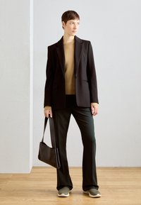 Svart blazer over en brun genser, kombinert med mørkegrønne skreddersydde bukser og olivengrønne sneakers. Holder en svart håndveske. Tregulv og lys grå bakgrunn.