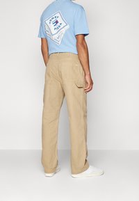 Jasnoniebieska bawełniana koszulka z grafiką pizzy na plecach, zestawiona z beżowymi spodniami typu cargo. Białe trampki dopełniają strój.