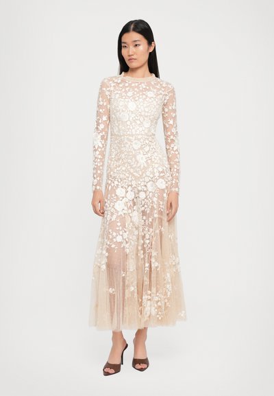Vrouw in een beige lange mouw tulle jurk met witte bloemenborduring en bruine open teen hakken, staand tegen een eenvoudige witte achtergrond.