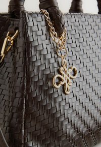 Coin d'un sac à main en cuir tressé noir avec anses tressées et chaîne en or ornée d'un charm ornemental complexe.