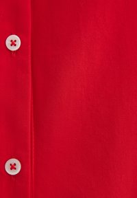 Roter Stoff mit glatter Textur, versehen mit einem weißen Knopf im Vierlochdesign, der entlang der Knopfleiste genäht ist.