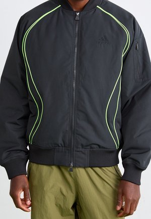 Schwarze Adidas Bomberjacke mit neon-grünem Paspel, Frontreißverschluss, gerippten Bündchen und Saum, getragen mit olivgrüner Hose.